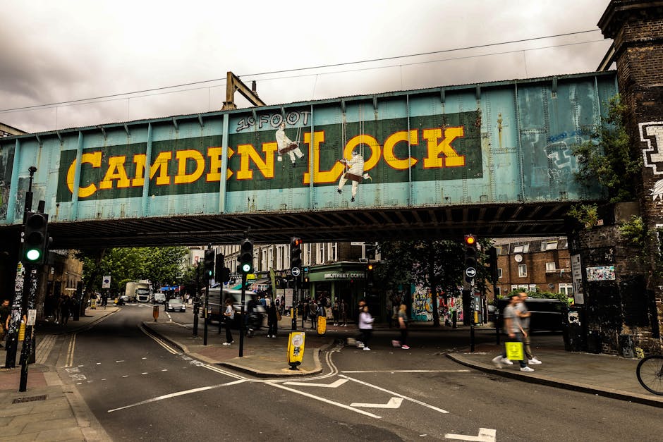 The image depicts the entrance to Camden Market in Camden, London, with a large arched sign reading 'CAMDEN MARKET' mounted on two metal poles. The scene is set on a pedestrian street lined with vario