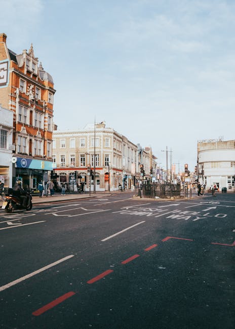 A wide view of Camden High Street during daytime showing a busy urban scene with historic and modern buildings lining both sides of the street. The buildings include a mix of brick and white facades, some with decorative architectural details, and retail shopfronts. There are pedestrians walking on the sidewalks, some waiting at street crossings or standing near shop entrances. The road has multiple lanes marked with white and red dashed lines, and a designated loading zone with clear signage. An electric scooter is parked on the pavement near the curb, and street lamps and traffic signals are visible further down the street. The sky is partly cloudy with soft sunlight illuminating the scene. This image relates to house removals and moving services such as those offered by Man with Van Camden, illustrating the urban environment where furniture transport, packing, and home relocation logistics often take place.