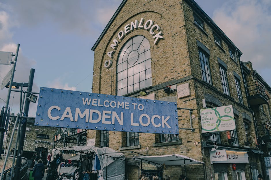 A wide view of Camden High Street during daytime showing a busy urban scene with historic and modern buildings lining both sides of the street. The buildings include a mix of brick and white facades, some with decorative architectural details, and retail shopfronts. There are pedestrians walking on the sidewalks, some waiting at street crossings or standing near shop entrances. The road has multiple lanes marked with white and red dashed lines, and a designated loading zone with clear signage. An electric scooter is parked on the pavement near the curb, and street lamps and traffic signals are visible further down the street. The sky is partly cloudy with soft sunlight illuminating the scene. This image relates to house removals and moving services such as those offered by Man with Van Camden, illustrating the urban environment where furniture transport, packing, and home relocation logistics often take place.