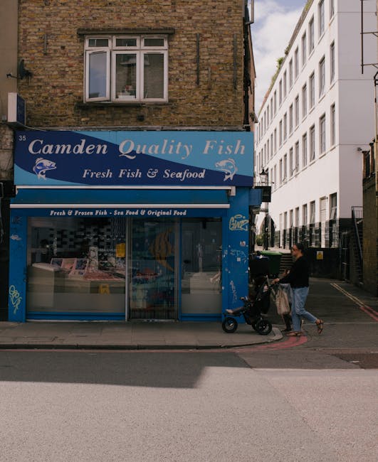 A street scene on Mornington Crescent in Camden, showing a mix of historical and modern buildings on either side of the road under a clear sky. The buildings include multi-storey shops and offices wit
